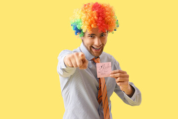 Businessman in clown wig with sticky paper pointing at viewer on yellow background. April Fools'...