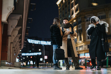 Three friends reunite on a cold urban evening, warmly dressed under city lights. A candid moment of urban life and friendship in wintertime. - obrazy, fototapety, plakaty