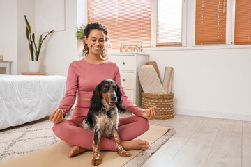Sporty young African-American woman with cocker spaniel meditating in bedroom