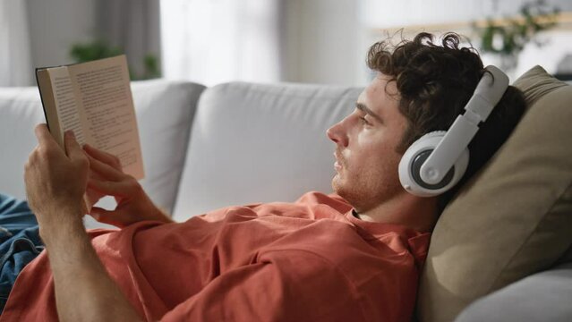 Headphones man reading book chilling sofa at home closeup. Guy laying couch