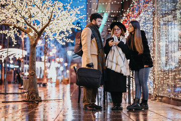 A group of young people engaged in a discussion on a city street adorned with twinkling festive...