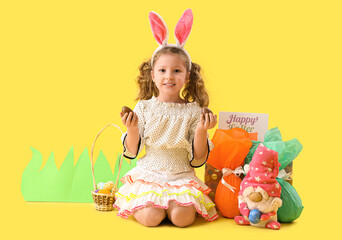 Cute little girl in bunny ears with chocolate Easter eggs and decor sitting on yellow background