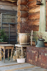 In the corner of a log house, a composition of old rustic items. A wooden bucket with branches, a green teapot, baskets with dry grass, a colored forged chest. Decoration.