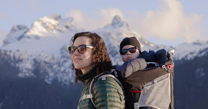 Happy Mother with Baby Boy in a Hiking Backpack Carrier with Mountains in the Background. Sunset sky. Squamish, BC Canada. Slow motion.