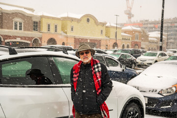 green economy: a senior citizen ( man with hat, beard, colorful carf and sunglasses) stands beside a new white electric vehicle in mall parking lot in a winter snowstorm