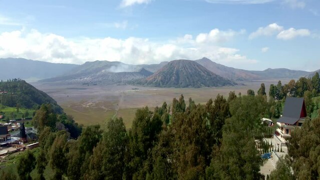 Capture the serene beauty of village life nestled in the foothills of Mount Bromo, as the majestic mountain provides a breathtaking backdrop, aerial 4k drone footage