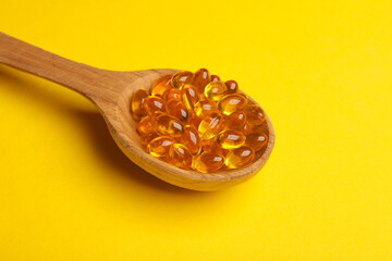 Fish oil capsules in wooden spoon on yellow background.