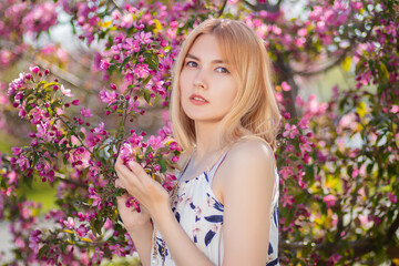 Fototapeta premium Portrait of an attractive young woman dressed in a dress posing against a background of blooming pink apple flowers. 