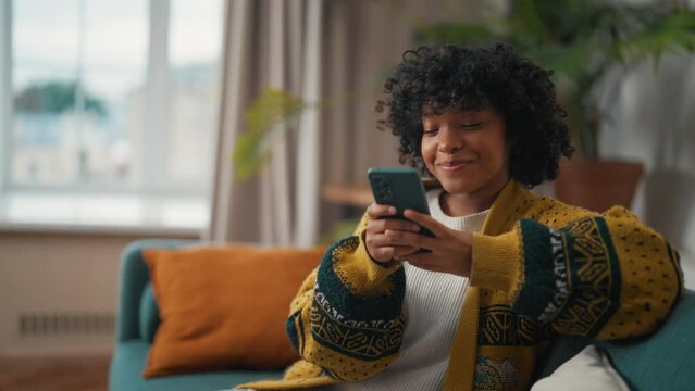 Young woman sitting with phone. African American woman hold in hand use mobile phone typing messages. Girl spend free spare time in living room. Communication on social networks, rest concept.
