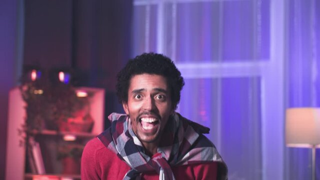 black male fan watches a live broadcast of a sports match and emotionally shouts hurray in a cozy room, looking at the camera