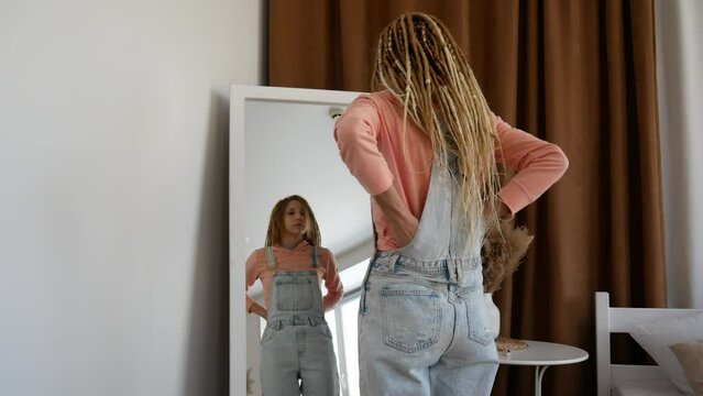 A beautiful girl with dreadlocks puts on denim overalls in front of a mirror