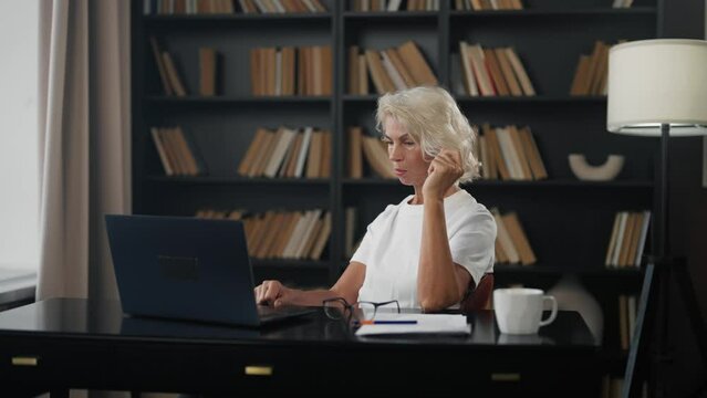 Senior woman freelancer works on laptop. Mature gray haired female specialist in glasses working at home office writing notes typing on computer. Expert doing her job. Freelance online distant work.
