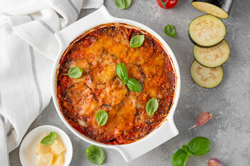 Eggplant casserole with cheese and tomato sauce in a white baking dish on a gray concrete background with ingredients for cooking. Vegetarian healthy food.