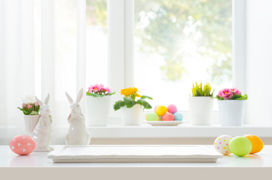 Easter time blank podium on table decorated with eggs, rabbit figures and blurred window flowers background