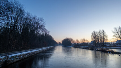 This image presents a tranquil winter morning by a canal, captured at the moment when the day is just beginning to break. The canal's surface is partially frozen, reflecting the cool tones of the sky