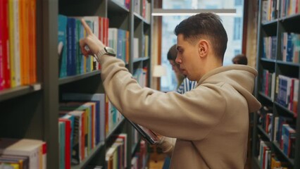 Young man in beige hoodie reaching for book on high library shelf, tablet in hand, with other students in blurred background - Powered by Adobe