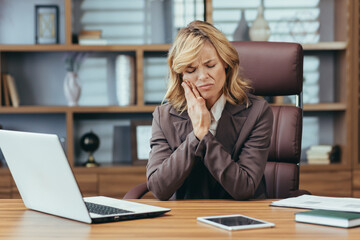 Mature businesswoman in pain, holding jaw suffering from toothache at office, showcasing workplace health issues.