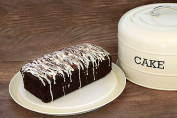 Chocolate chip cake with icing drizzle on metal cream tray with lid on rustic wood background....