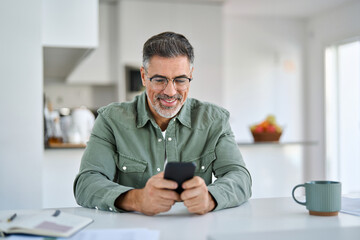 Happy old middle aged man using mobile phone sitting at kitchen table. Smiling senior mature...