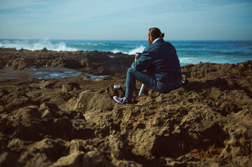 Mature Caucasian bearded man taking coffee break while hiking in the rocky beach, sitting relaxed on the rocky cliff