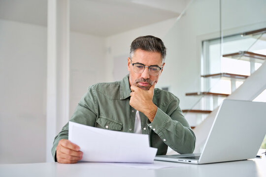 Serious middle aged mature man entrepreneur wearing eyeglasses looking at paper bill checking insurance invoice or tax document calculating expenses working at home using laptop computer. - Powered by Adobe