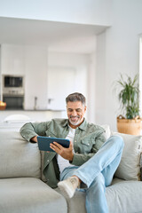 Smiling middle aged man using digital tablet relaxing on couch at home. Happy mature male user...