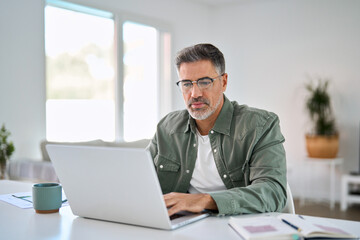 Serious older mature middle aged man wearing eyeglasses looking at computer technology sitting at...
