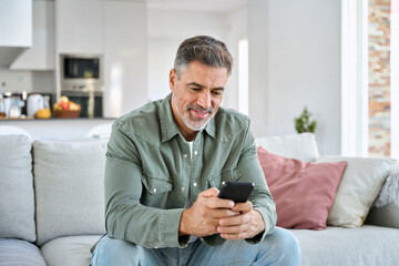 Middle aged old man using smartphone relaxing on couch at home. Happy senior mature male user holding cellphone browsing internet, texting messages on mobile cell phone technology sitting on sofa. - obrazy, fototapety, plakaty