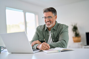 Happy smiling middle aged older mature man wearing glasses looking at laptop using computer writing notes watching webinar sitting at home table. Hybrid work, elearning, online learning concept. - Powered by Adobe