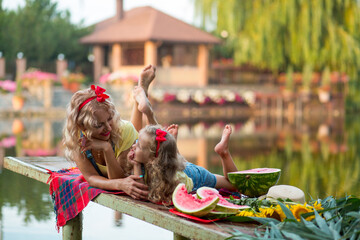 Blonde mother and three-year-old daughter in denim shorts with straps with red bows eat watermelon...