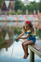 A three-year-old blond girl in denim shorts with straps sits on a pier and pours water on her bare...