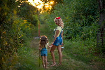 Two blond barefoot fisherwomen in denim shorts with straps with a fishing rod and bucket on a road...