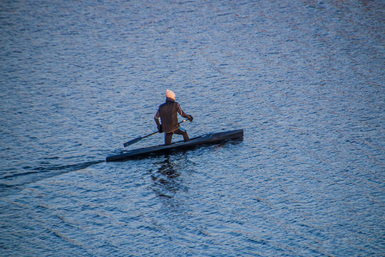 An athlete with a black oar in his hands swims in the lake at sunset