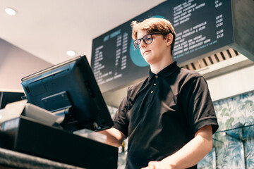 Young waiter serving customer at cash point in cafe. Man working with POS terminal. Cashier, barista checking for payment receipt. Hospitality, server and preparing a slip at the till in coffee shop - obrazy, fototapety, plakaty