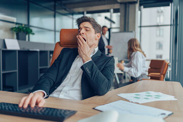 Young overtired businessman yawning while working inside modern office with computer