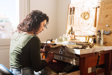 Young female jeweller sitting at table while making jewellery in workshop