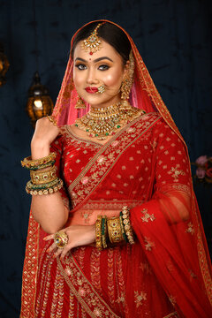 Stunning Indian bride dressed in traditional red bridal lehenga with heavy gold jewellery and veil smiles tenderly in studio lighting. Wedding fashion and lifestyle.