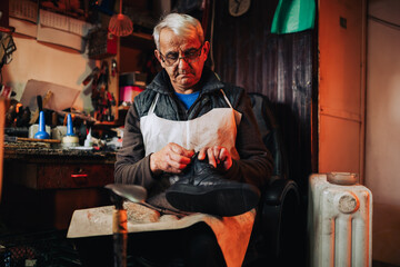 A senior cobbler is sitting at workshop and examining boots.