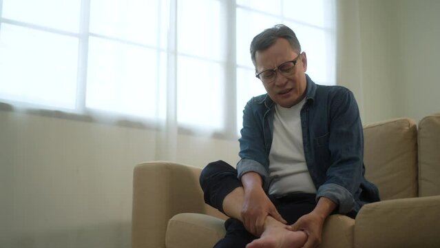 Senior man suffering from foot pain when sitting on sofa, toe and ankle problem in retirement, unhappy older man with chronic joint disease at home, leg arthritis and health care