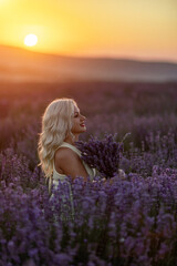 Fototapeta na wymiar Blonde woman poses in lavender field at sunset. Happy woman in white dress holds lavender bouquet. Aromatherapy concept, lavender oil, photo session in lavender