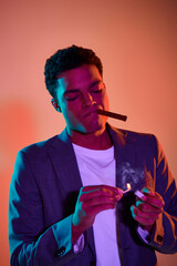 african american guy with cigar holding match with fire on pink background with blue lighting