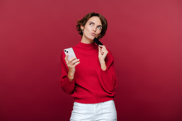 Beautiful pensive woman holding mobile phone and credit card, thinking what to buy
