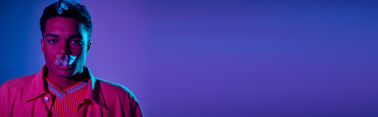 young african american man exhaling smoke against a blue background with purple lighting, banner