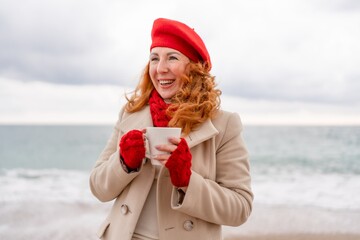 Obraz premium Woman beach in red beret scarf and mitts holding a cup of tea in his hands. Depicting beach relaxation and cozy attire. Walks by the sea