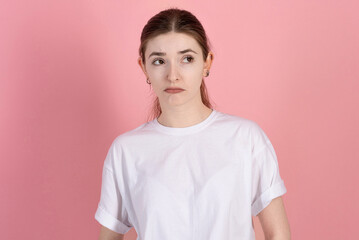Fototapeta premium Portrait of a sad and depressed attractive young brunette woman wearing a casual white t-shirt, sulking and frowning isolated on a pink studio background.