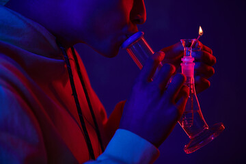 cropped african american man in hoodie lighting glass bong on dark blue background with red light