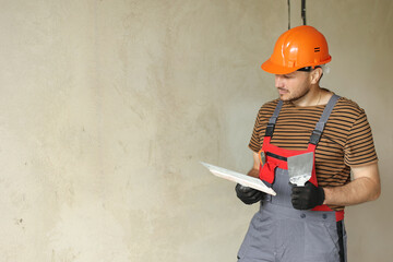 repairman in overalls and protective orange helmet holds two spatulas for plastering concrete wall...