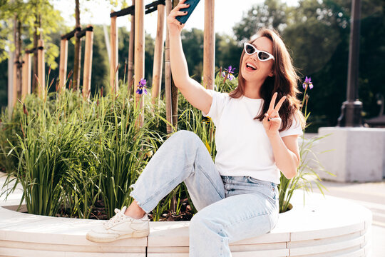 Young beautiful smiling hipster woman in trendy summer white t-shirt and jeans clothes. Sexy carefree model posing in the street at sunset. Positive model outdoors. In sunglasses. Takes selfie photos