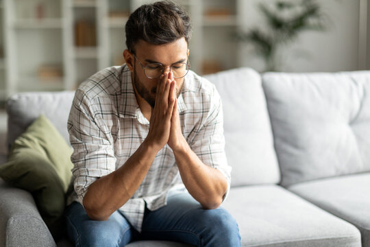 Mental health, depression concept. Frustrated young indian guy sitting on sofa alone, looking down, have hard thoughts, copy space