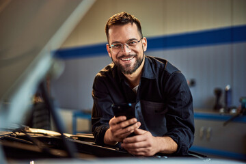 A portrait of a smiling mechanic man, leaning on the car, using a mobile phone.